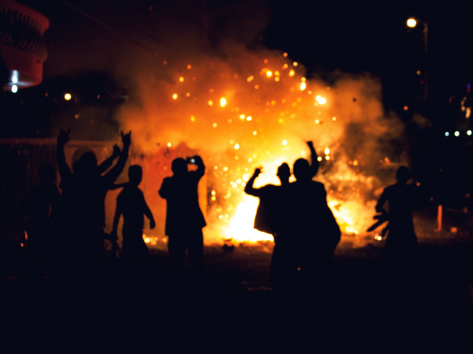 Photo of people gathered round a fire with scattering embers, arms raised.